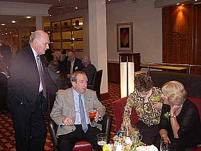 74th meet in the lounge. A lounge bar with people sitting and standing around a table.